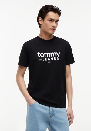 Bărbat tânăr purtând un tricou negru "Tommy Jeans" și blugi albastre deschis, stând cu o mână în buzunar, pe un fundal alb simplu.