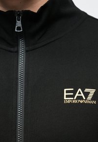 Čierna bunda na zips s vysokým golierom, štruktúrovanou tkaninou a zlatým logom EA7 na ľavej strane hrudníka. Zips je viditeľný.