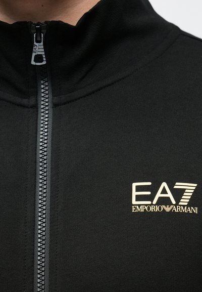 Черно яке с цип и висока яка, изработено от текстурирана материя, с златно лого EA7 на лявата част на гърдите. Зъбите на ципа са видими.