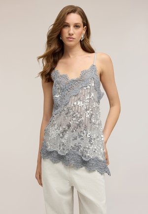 Kvinde med langt brunt hår iført sølv glimmer blonde-trimmet camisole og hvide bukser, stående mod en neutral baggrund.