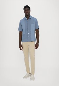 Blaues kurzärmliges Hemd mit einer großen aufgesetzten Tasche; kombiniert mit hellbeigen Slim-Fit-Hosen und weißen Sneakers. Einfarbiger Hintergrund.