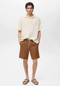 Beige pletené polo tričko s krátkým rukávem a límečkem, doplněné hnědými šortkami s elastickým pasem a šňůrkou. Celkový vzhled doplňují hnědé sandály.