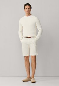 Hombre de pie con las manos en los bolsillos, lleva un suéter de punto color crema, pantalones cortos beige con cinturón marrón y zapatos beige sin cordones, sobre un fondo liso.