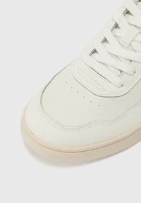 Gros plan sur l'avant d'une basket blanche en cuir perforé, avec des lacets blancs et une semelle en caoutchouc beige clair sur un fond uni.