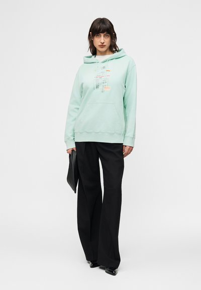 Γυναίκα που φοράει φούτερ μέντας με σχέδιο εκτυπωμένο, μαύρο παντελόνι φαρδύ, μαύρα παπούτσια και κρατάει ένα μαύρο τσαντάκι, με φόντο λευκό.