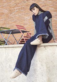 Wide-leg dark denim pants with frayed hems, paired with a black and white striped turtleneck and beige slip-on shoes, against a brick backdrop.