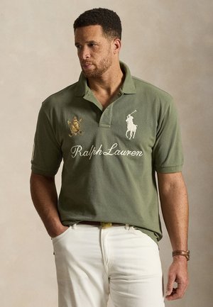 Άνδρας που φοράει πράσινο χακί πόλο Ralph Lauren με λογότυπα και λευκό παντελόνι, στέκεται με το αριστερό χέρι στην τσέπη μπροστά σε μπεζ φόντο.