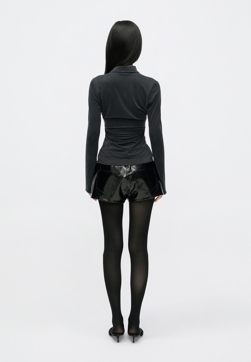 Sort langærmet, tætsiddende top, parret med skinnende sorte shorts. Modellen bærer sorte strømpebukser og sko med høje hæle. Glatte teksturer og tætsiddende design.