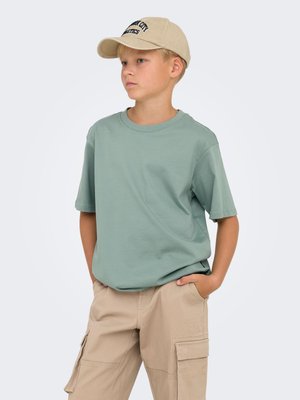 Lichtgroene katoenen t-shirt met korte mouwen, gecombineerd met beige cargobroek met zijzakken; draagt een beige pet met zwarte tekst.