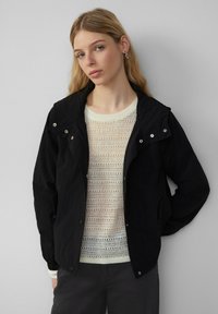Veste zippée noire avec capuche, dotée de boutons-pression argentés, associée à un pull en tricot crème avec un motif en dentelle.