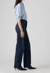 Kvinde iført lyseblå, stribet skjorte, mørke jeans med vide ben og sorte hæle, stående mod en ensfarvet baggrund.