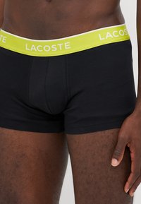 Čierne bavlnené boxerky s jasne žltým pásom s bielym nápisom "LACOSTE", s tesným strihom a hladkou textúrou.