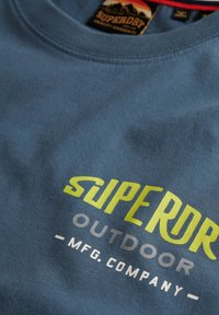 Camiseta de algodón azul con la inscripción "SUPERDRY" en texto amarillo brillante y gris, con un cuello redondo y una textura suave. Etiqueta colgante visible.