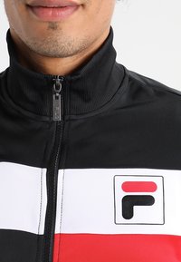 Sort zip-up jakke med høj krave, prydet med vandrette striber i hvid og rød, samt et Fila-logo på venstre bryst.