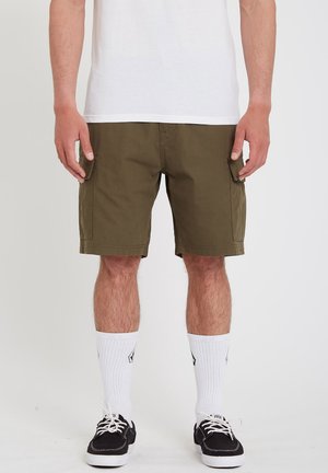 Mann trägt olivgrüne Cargoshorts, weiße Sneakersocken, schwarze Sneakers und ein weißes T-Shirt und steht vor einem schlichten Hintergrund.