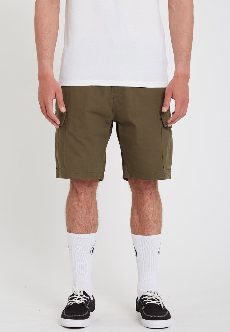 Homme portant un short cargo vert olive, des chaussettes blanches, des baskets noires et un t-shirt blanc, debout devant un fond uni.