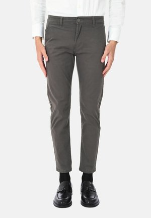 Pantaloni in cotone slim-fit verde oliva con design a cinque tasche, chiusura frontale con bottone e leggero elastico. Indossati con mocassini neri e calzini.