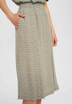 Femme portant une jupe bleu clair avec un motif tacheté marron, une main dans la poche latérale, l'autre pendant le long du corps.