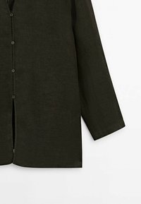 Chemise en lin vert foncé à manches longues, fermeture à boutons, et fentes latérales pour faciliter les mouvements. Le tissu texturé améliore la respirabilité.