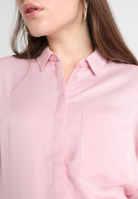 Light pink button-up shirt with a collar, single chest pocket, and smooth fabric texture, featuring subtle stitching and buttons.