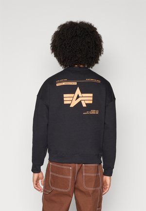 Osoba s kudrnatými vlasy, která má na sobě černou mikinu Alpha Industries s logem a nápisem, a hnědé kalhoty s bílým prošíváním, z profilu od zády.