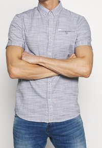 Camisa de manga corta a cuadros en azul claro y blanco. Con cuello abotonado, bolsillo frontal y tejido texturizado. Combinada con vaqueros azules.