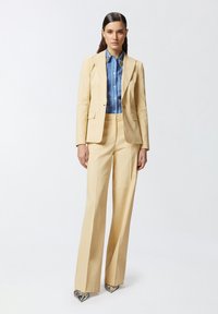 Completo giallo su misura con giacca aderente e pantaloni a gamba larga, abbinato a una camicia a righe blu. Scarpe con tacco alto a fantasia completano il look.