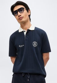 Άνδρας που φοράει μπλε σκούρο πόλο με λευκό γιακά και σκούρα γυαλιά ηλίου, στέκεται μπροστά σε απλό ανοιχτό φόντο.