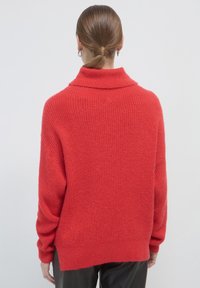 Frau mit zusammengebundenem Haar, die einen lockeren, leuchtend roten Strickpullover und schwarze Hosen trägt, steht mit dem Rücken zu einem schlichten weißen Hintergrund.