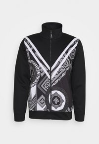 Svart zip-up jacka med hög krage, med vita och grå geometriska och blomster mönster som bildar en V-form fram.