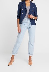 Sini sininen kaksirivinen bleiseri kultanappien kanssa, harmaa pitsi-toppi, vaaleansiniset suorat farkut ja vaaleanruskeat kärjelliset korkokengät.