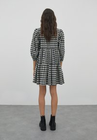 Mustavalkoinen ruudullinen mekko puffihihoilla, kerroksellisella helmalla ja rentolla istuvuudella. Yhdistetty mustiin nilkkabootseihin, nähty takaapäin.