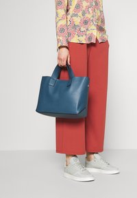 Modrá texturovaná kabelka s krátkými uchy, obdélníkový tvar. Nosičná s červenými širokými kalhotami a světle šedými teniskami. V pozadí květinová košile.