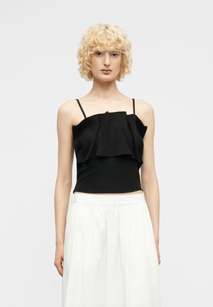 Femme blonde aux cheveux bouclés portant un haut noir sans manches à volants et une jupe blanche taille haute sur un fond gris uni.