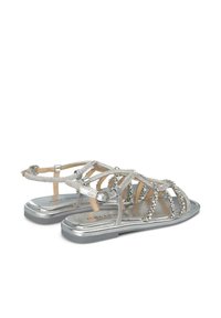 Sandalias planas plateadas con un diseño de tiras entrelazadas que presentan acentos metálicos y brillantes decorativos en la parte superior. Textura suave y ligeras.
