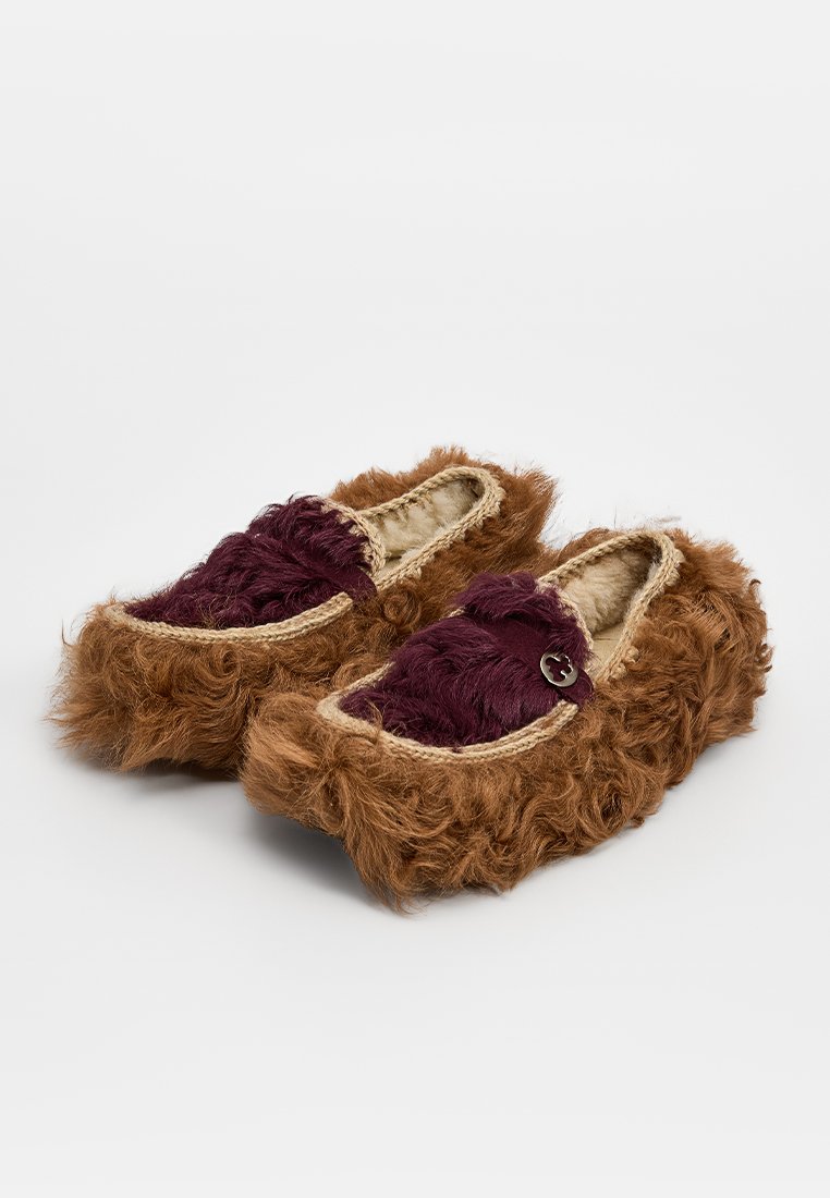 Braune Kunstfellslipper mit einem burgunderfarbenen Stoffeinsatz, geflochtenem Rand und dekorativen Knopfdetails. Weiches, strukturiertes, leicht anzuziehendendes Design.