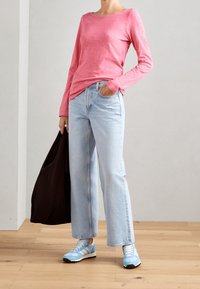 Rosa Langarmshirt, hellblaue weit geschnittene Jeans und blaue Sportschuhe. Schwarze Tote-Bag in der Hand, stehend auf einem Holzboden vor einer hellen Wand.