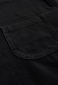 Tissu noir à texture lisse, avec une poche arrondie. Les coutures contrastées sont soigneusement finies autour des bords de la poche.