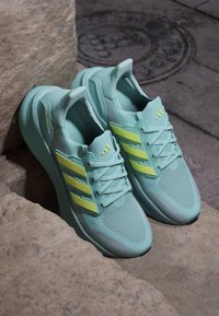 Helle blaue Laufschuhe mit einem atmungsaktiven Mesh-Obermaterial, drei gelben Streifen, Schnürsenkeln und gepolsterten Sohlen mit "ULTRAGOOSt"-Markierung.