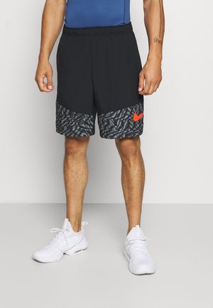 Mann trägt schwarze Sportshorts mit einem grauen gemusterten Band und rotem Nike-Logo, weiße Sneakers und ein blaues Oberteil, steht auf grauem Boden.