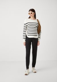 Stripet genser i svart og hvitt, med ribbestruktur, med knapper på siden; kombinert med svarte bukser og beige sneakers.