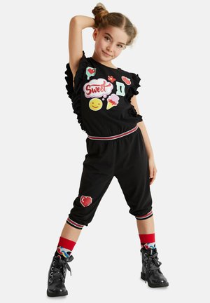 Schwarzes Zweiteiler-Outfit mit Rüschensleeves, ausgestattet mit bunten Applikationen und einem kontrastierenden gestreiften Bund. Kombiniert mit schwarzen Stiefeln und gemusterten Socken.