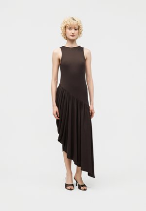 Kobieta o krótkich, kręconych, blond włosach, ubrana w bezrękawnikową, asymetryczną, ciemnobrązową sukienkę oraz czarne sandały na obcasie z odkrytymi palcami, stojąca na jednolitym tle.