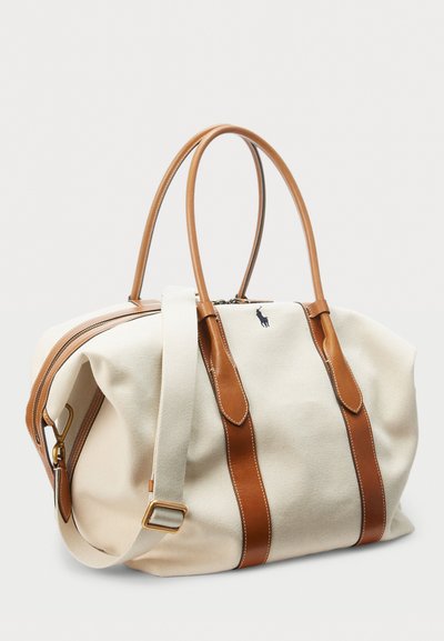 Canvas-Reisetasche mit braunen Ledergriffen und Akzenten, ausgestattet mit einem Reißverschluss und einem abnehmbaren Riemen. Gesticktes Logo an der Seite.