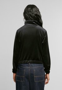 Svart velurjakke med høy krage, elastisk midje og mansjetter. Bakdelen har en jevn tekstur og en tettsittende design. Dongeribukser er synlige.