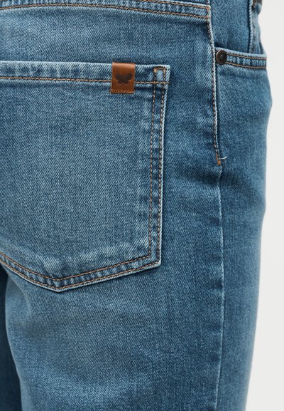 Κοντινό πλάνο πίσω τσέπης μπλε τζιν με πορτοκαλί ραφές και ένα μικρό καφέ δερμάτινο ταμπελάκι με λογότυπο πεταλούδας.