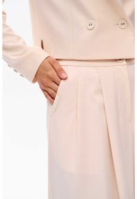 Lichtbeige tailsuit met een dubbelgeknoopt jasje, gladde textuur, knoopdetails en een label op de zak van de broek.