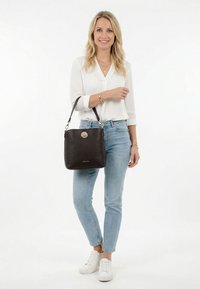 Žena v bílé blůze a džínách se usmívá, drží tmavě hnědou koženou kabelku s popruhem přes rameno.