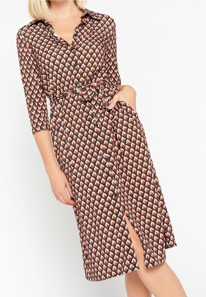 Kobieta w zapinanej na guziki, przewiązanej paskiem sukience midi z geometrycznym wzorem w odcieniach brązu, beżu i czerni, z jedną ręką w kieszeni, z rozcięciem z przodu.