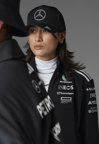 Schwarze Mütze mit silbernem Mercedes-Benz-Logo, kombiniert mit einer schwarzen Zip-Jacke, die mehrere Sponsorenlogos in Weiß trägt. Darunter ein weißer Rollkragenpullover.